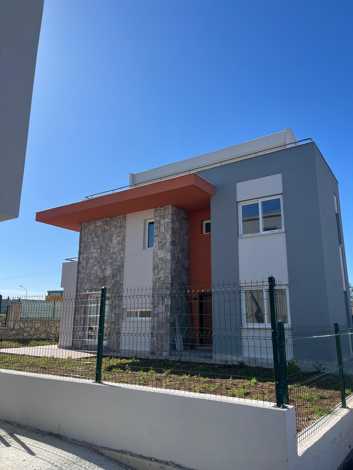 Modern two-story house with gray facade, orange cantilevered balcony, stone column, and white window trim behind a green metal fence in a sunny setting.
