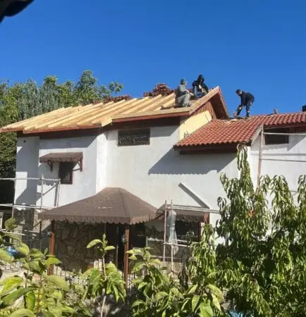 Construction workers on a pitched roof installing wooden rafters on a sunny day, a white house beneath with scaffolding nearby and green trees around.